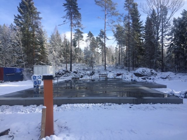 Nybyggd betonggrund i snöig terräng med skog i bakgrunden och byggmaterial i förgrunden.