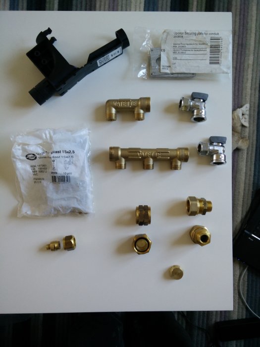 Olika VVS-detaljer för renovering, inklusive mässingskopplingar och säkringsplatta, uppställda på en vit yta.