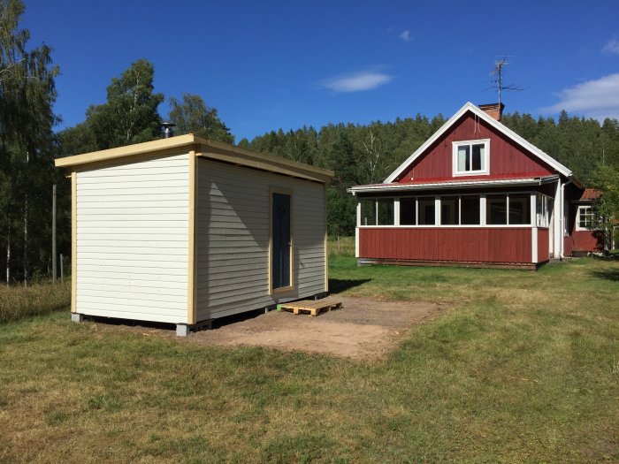 Ommålad bastubyggnad i trä bredvid äldre rött hus med vita knutar på en gräsmark.