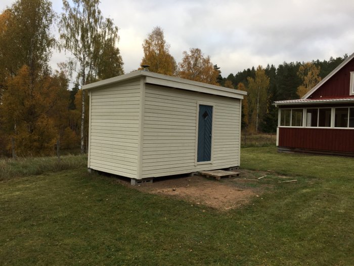 Nybyggt, omålat utvändigt bastuhus med blå dörr på gården omgiven av höstträd.