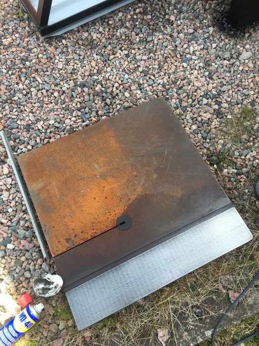 Rostig metallplatta på mark med småsten och WD-40 sprayburk i förgrunden.