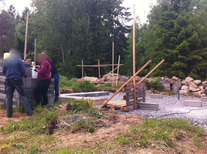 Två personer arbetar vid grundläggningsarbete i skogsmiljö; träpålar, gråsten, betongblock synliga.