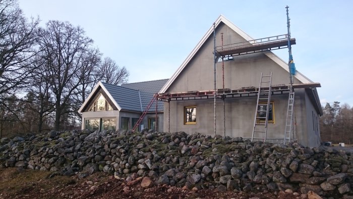 Hus under renovering med pågående putsarbete, byggställning och stenmur i förgrunden.