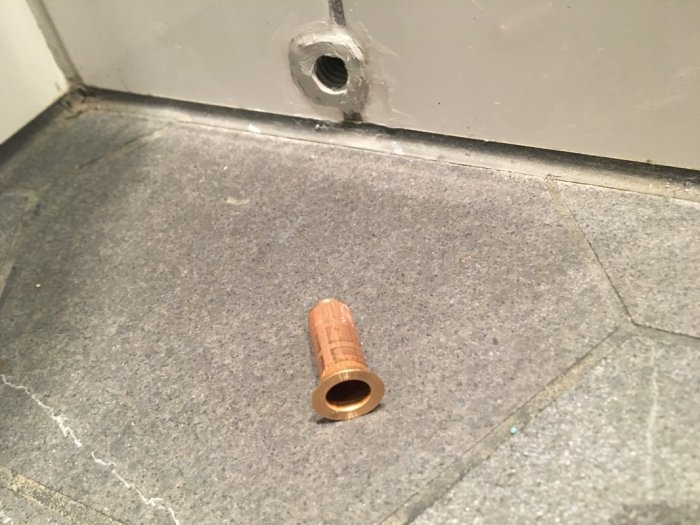Mässingsfärgad ventil på ett grått kakelgolv bredvid ett skvallerrör med våtrumssilikon runt kanten.