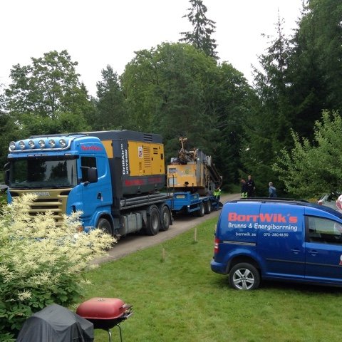 Brunnsborrningsmaskin på lastbil och servicebil på trång grusväg med grönska.