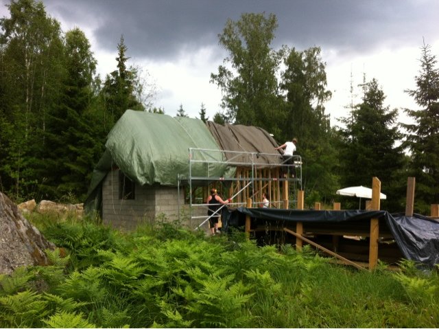 Ett pågående byggprojekt med hus under konstruktion, täckt av en presenning, med arbetare på taket och i trädgården.