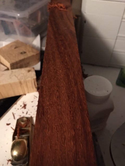 Hyvlad kant på mörkt träplank med synlig ådring, eventuellt mahogny, på ett rörigt arbetsbord.