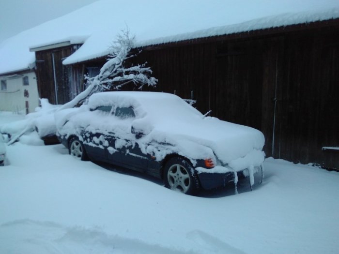 Bil täckt med tjockt lager snö framför en lada, vilket visar nattens kraftiga snöfall.