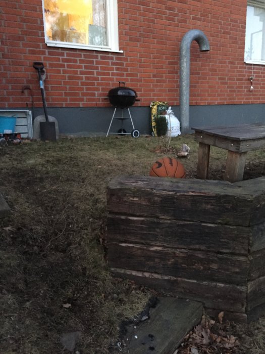 Framtidig plats för trall, nära husvägg med fönster, grill, spade och pall på gräs intill träkuber och basketboll.