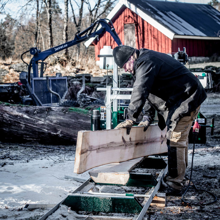 Person som sågar askplank på mobilt sågverk utomhus med röd lada i bakgrunden.