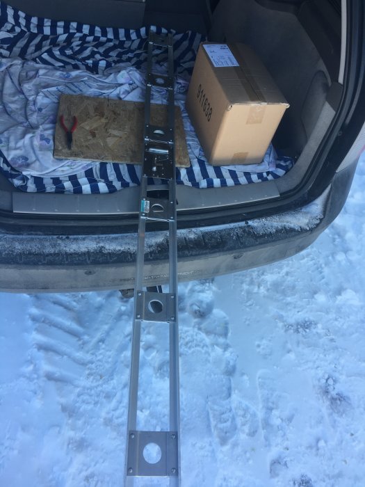 Metallstege i en bilbagage med ett paket och en skruvmejsel på en snötäckt mark.