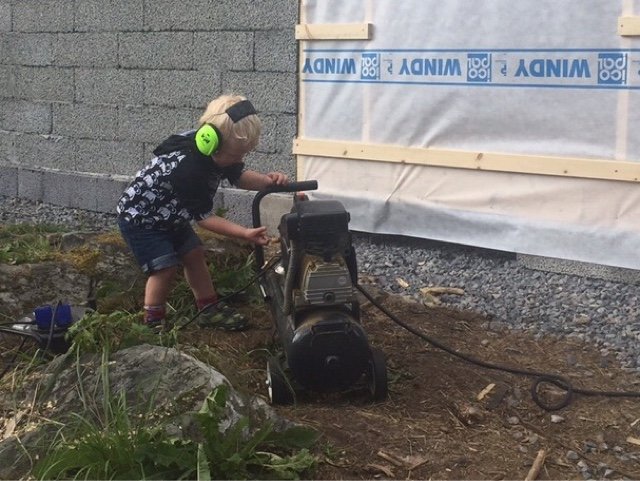 Barn med hörselskydd leker med en utomhuskompressor vid ett hus under konstruktion.