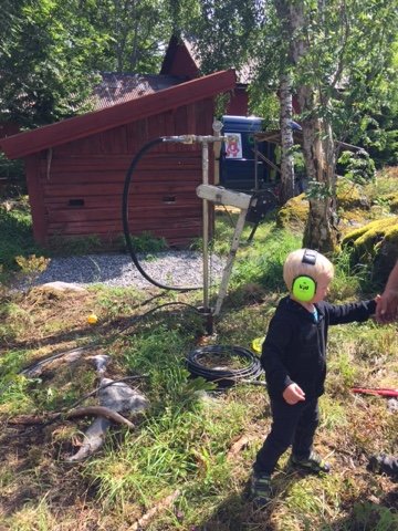 Ett barn i hjälm vid en brunnsborrning med en pump och timer, med ett rött hus och grönska i bakgrunden.