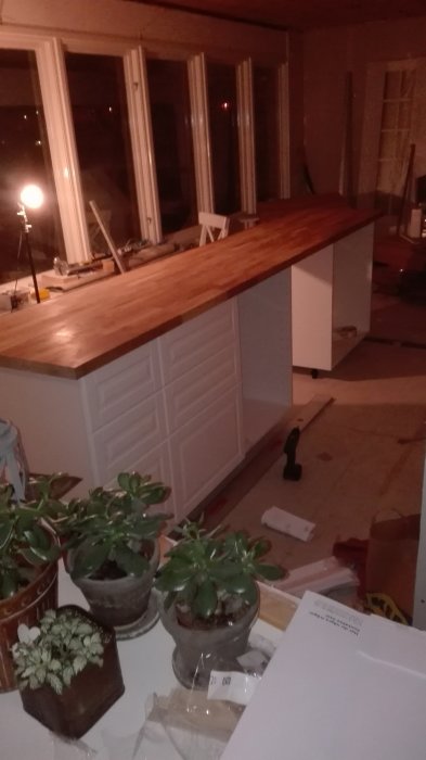 Under konstruktion köksö med träbänkskiva, vita skåpsluckor och verktyg omkring i kvällsbelysning.
