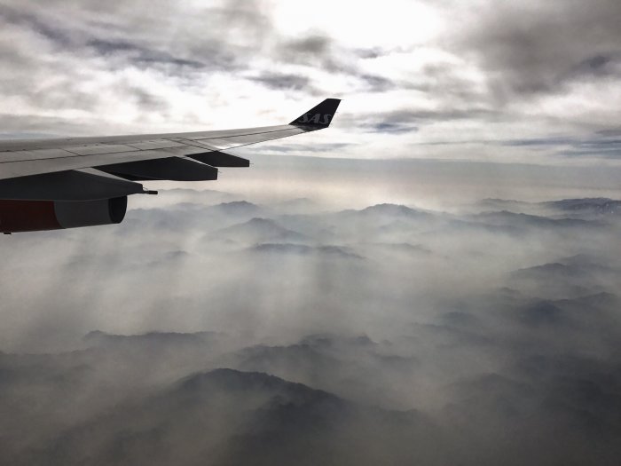 Flygplansvinge ovan moln och bergstoppar, dimmig vy, resefoto, lugn atmosfär, gråblå toner.