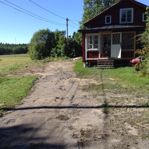 Grävd upp gräsmatta framför röd stuga med vit veranda, grusväg och strömkabel ovanför.