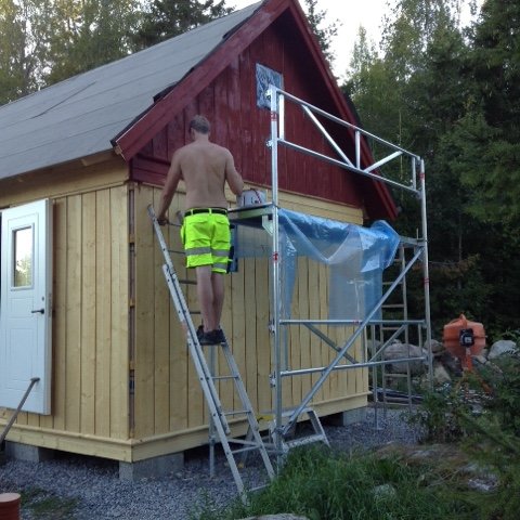 Man i arbetskläder på en stege vid en nypanelad stuga som målas faluröd, med byggmaterial i bakgrunden.