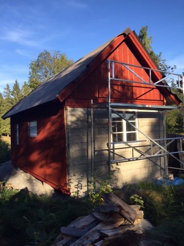 En rödmålad stuga under renovering med nya panelbrädor och byggnadsställningar runt fasaden.