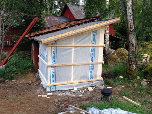 Isolerat pumphus under konstruktion med isolermaterial och träreglar synliga, intill skog och grannbyggnad.