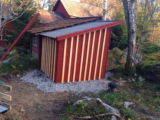 Nybyggt isolerat pumphus med vertikal röd och gul panel intill ett äldre rött hus med vita knutar på en grusgrund.