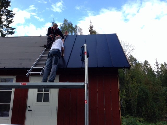 Två personer installerar svart Plannja Trend plåttak på en röd byggnad.