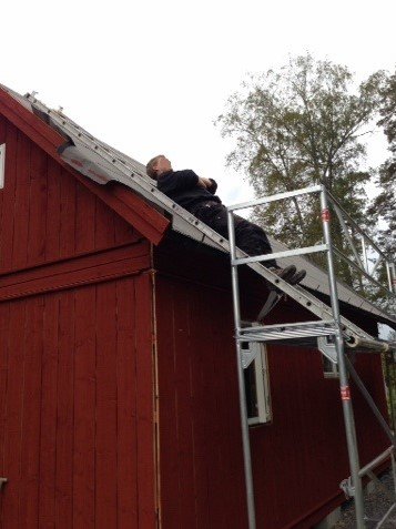 Person på en byggställning som arbetar med att montera svart plåttak på en röd träbyggnad.