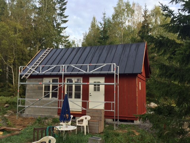 Rött hus med nyinstallerat svart plåttak och byggställningar runtom, omgiven av skog.