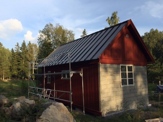 Röd trästuga med nytt svart plåttak och byggställning runt huset, omgiven av naturen i soligt väder.