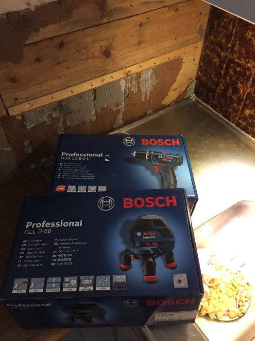 Bosch skruvdragare och lasernivå i förpackningar framför en rivet kakelvägg.