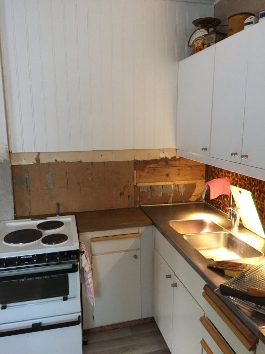 Kök under renovering med borttaget kakel och synliga väggskivor bakom vit spis och diskbänk.