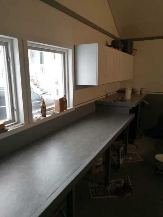 Nybyggd arbetsbänk i ett rum med fönster och vita skåp, förberedd för lådor och LED-belysning.