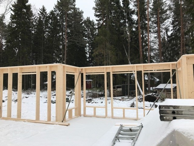 Trästomme till ett hus under uppbyggnad i ett snöigt landskap med träd i bakgrunden.