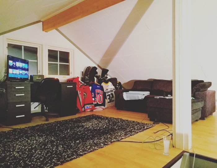 Provisorisk soffa och skrivbord på ett loft med snedtak och idrottsutrustning vid fönstret.