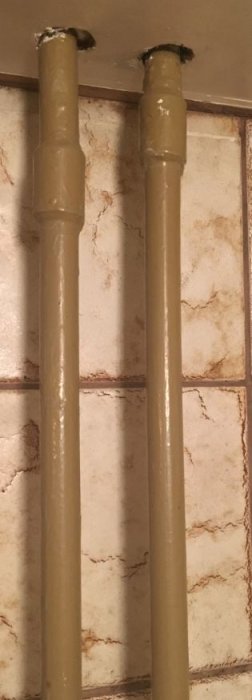 Två stålrör för element som kommer ut ur en vägg, målade beige mot kakelbakgrund.