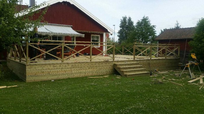 Nästan färdigbyggd träaltan med räcke vid röd stuga, verktyg och material syns på gräsmatta.