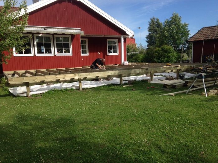 En person bygger en träaltan vid ett rött hus omgiven av grönt gräs.