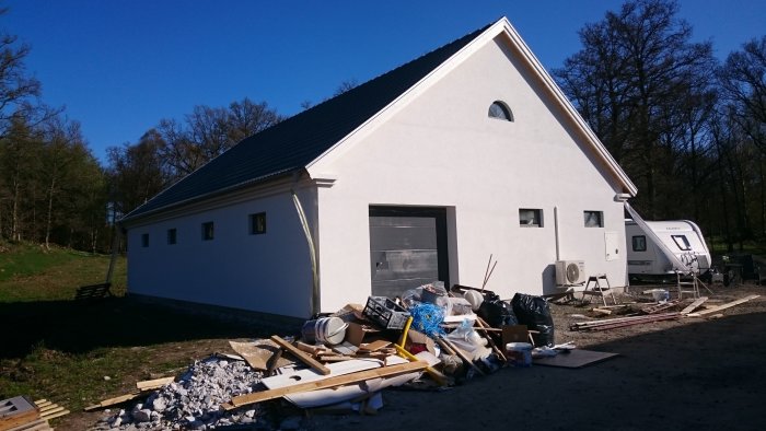 Vitt nyputsat hus med grå takpannor, byggavfall och husvagn bredvid under en klarblå himmel.