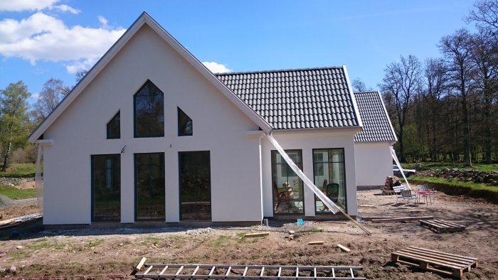 Vitputsad nybyggd villa i skånsk stil, med svarta fönsterkarmar, grå takpannor och provisoriska stuprör, under en klarblå himmel.