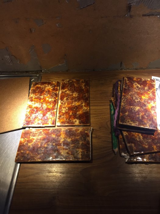 Sex keramiska plattor med rostliknande mönster på ett träbord, vissa skadade, nära en vägg med avflagande färg.