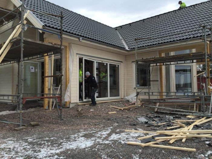 Hantverkare monterar vikparti i tegelklädd husfasad med byggställningar och materialrester på marken.