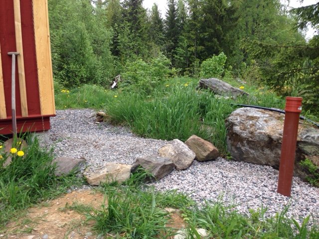 Nygrusat hörn med stenar bredvid en röd stuga, stor hörnsten och gräsmark med blommor i bakgrunden.