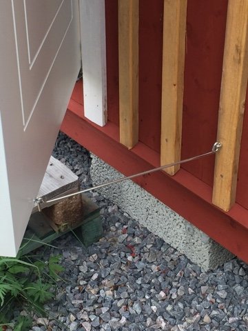 Hörn av hus med nyinstallerad hasp på rödlackerad dörr, grus och stenar vid grunden.