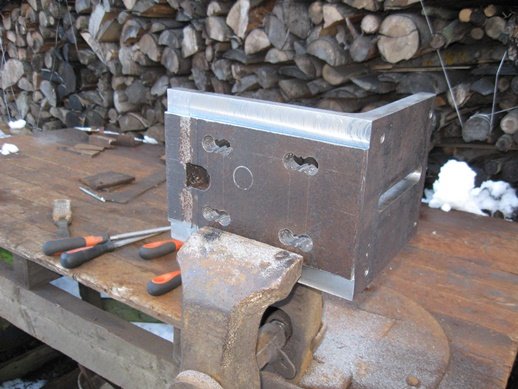 Metallblock med avlånga borrhål och filverktyg på arbetsbänk framför vedstapel.