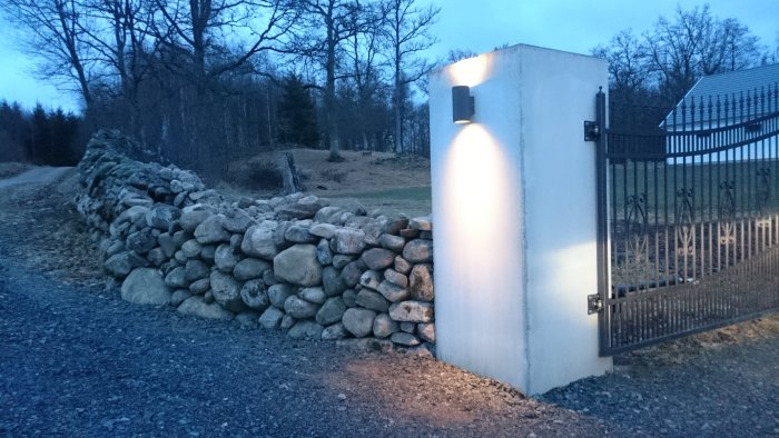 Stenmur längs en grusväg med lykta på en vit pelare vid en grind.