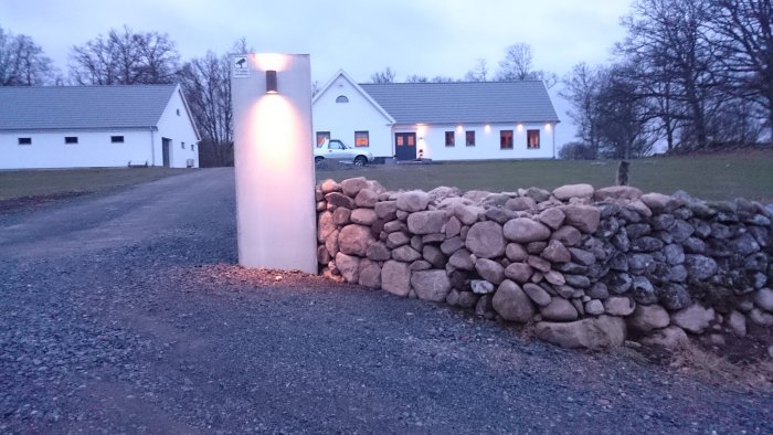 Vitputsat hus med garage och belysta grindstolpar vid en stenmur längs en grusväg i skymningen.