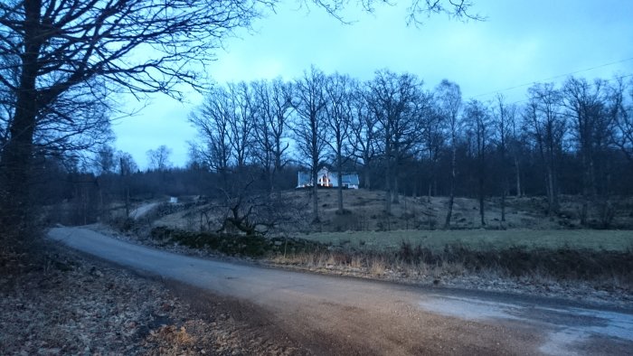 Hus på en kulle med tänt ljus i fönstren, sjön vid skymning, omgivet av träd och en grusväg i förgrunden.