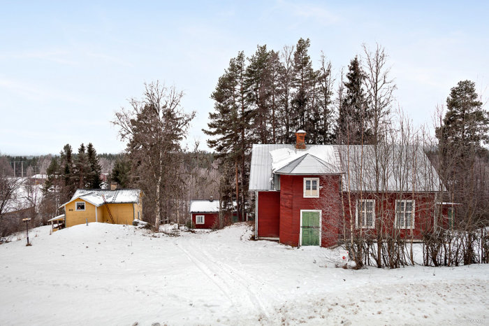 Äldre rött torp med vita knutar omgivet av snö, träd och andra små byggnader i landsbygdsmiljö.