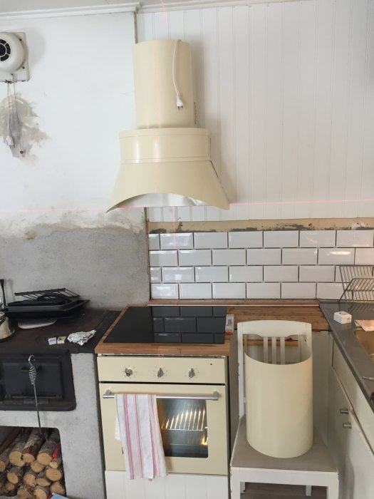 Ett kök under renovering med vit spis och fläkt, nyinstallerade vita kakelplattor och oskyddad vägg.