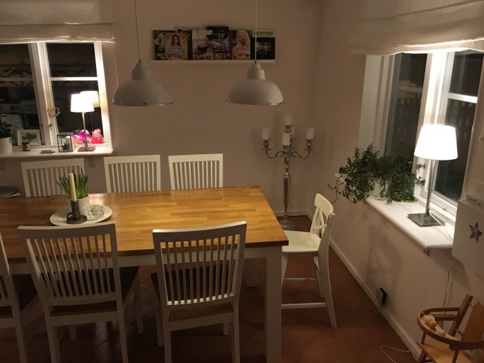 Kvällsbild av ett matrum före renovering, med ett träbord, vita stolar, fönster med blommor och taklampor.