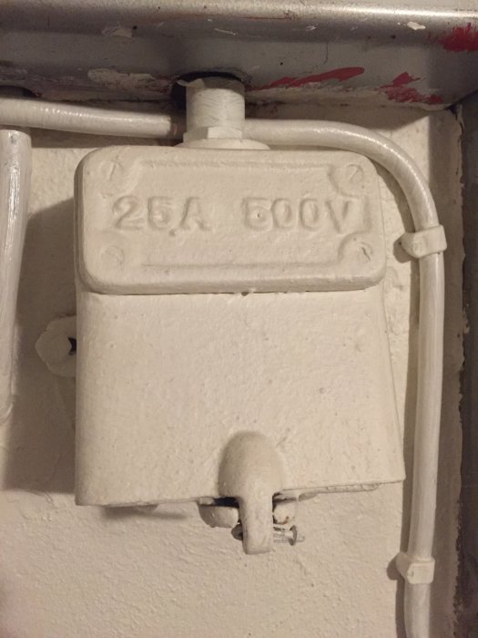 Gammalt vitmålat uttag märkt med "25A 500V" i en byggnad, omgivet av rör och kablar.